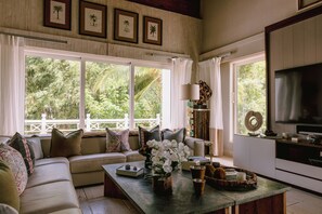Sala de estar con sofá, una mesa de centro y televisor. Se ven cuadros en la pared y vistas de vegetación al exterior.