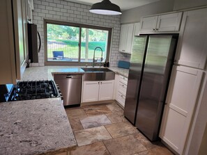 A kitchen with a gas stove, stainless steel appliances, a window with a view of greenery, and white cabinetry. - Beautiful home on the 8th hole of LV Country Club! (Las Vegas)