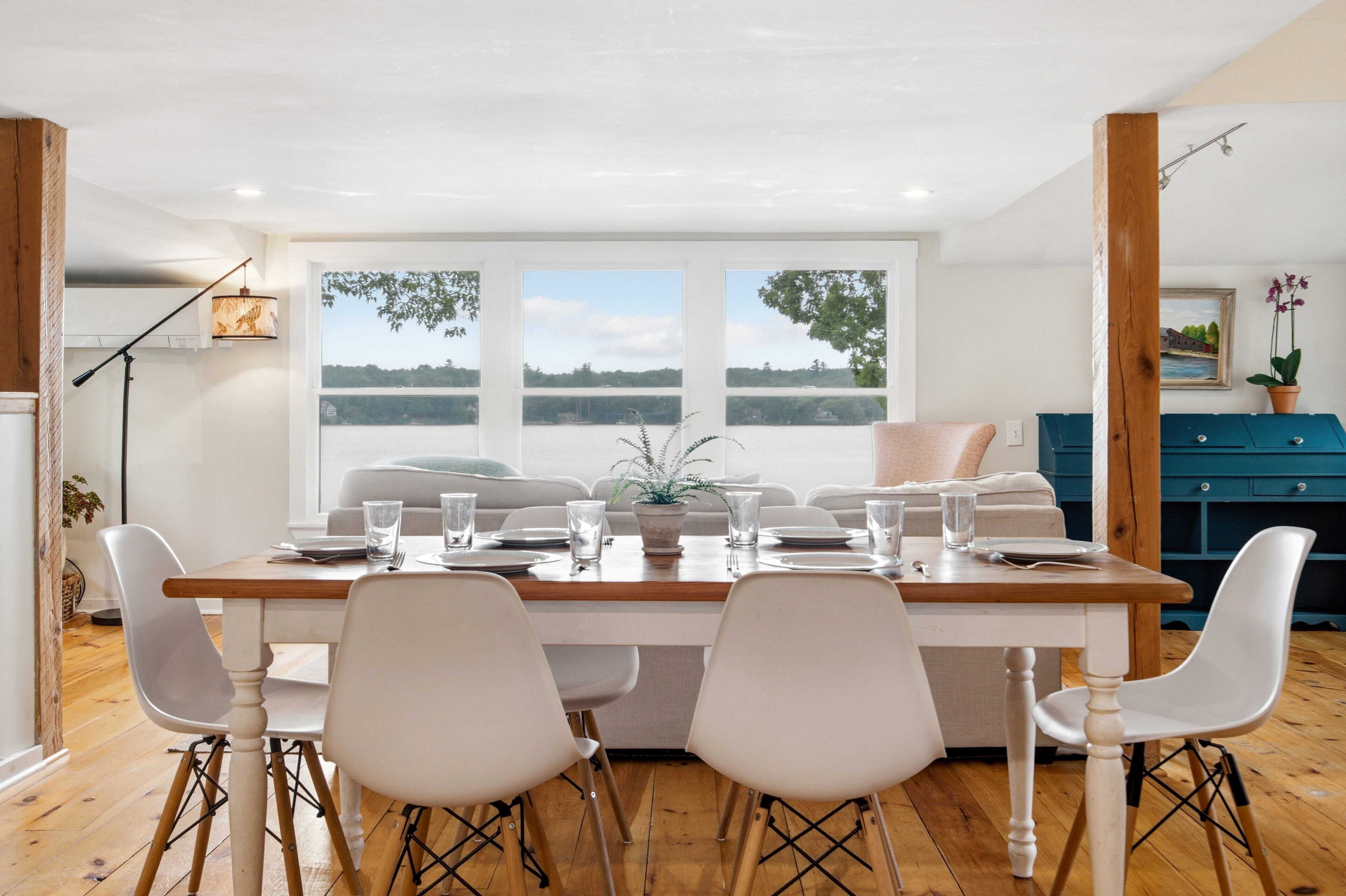 A dining area with a wooden table, white chairs, and a view of a lake through large windows.