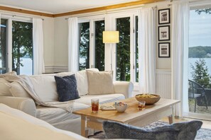 A living room with a sofa, a coffee table, and a view of a lake through large windows. - Highland Lake Cottage/Waterfront/Dock/Kayaks (Windham)