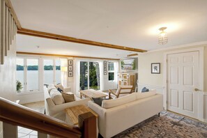 A spacious living area with a large window overlooking a body of water, a wooden staircase, and a view of the outdoors. - Highland Lake Cottage/Waterfront/Dock/Kayaks (Windham)