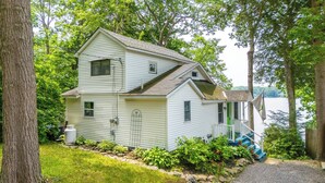 A two-story house with a porch, surrounded by trees and a body of water. - Highland Lake Cottage/Waterfront/Dock/Kayaks (Windham)