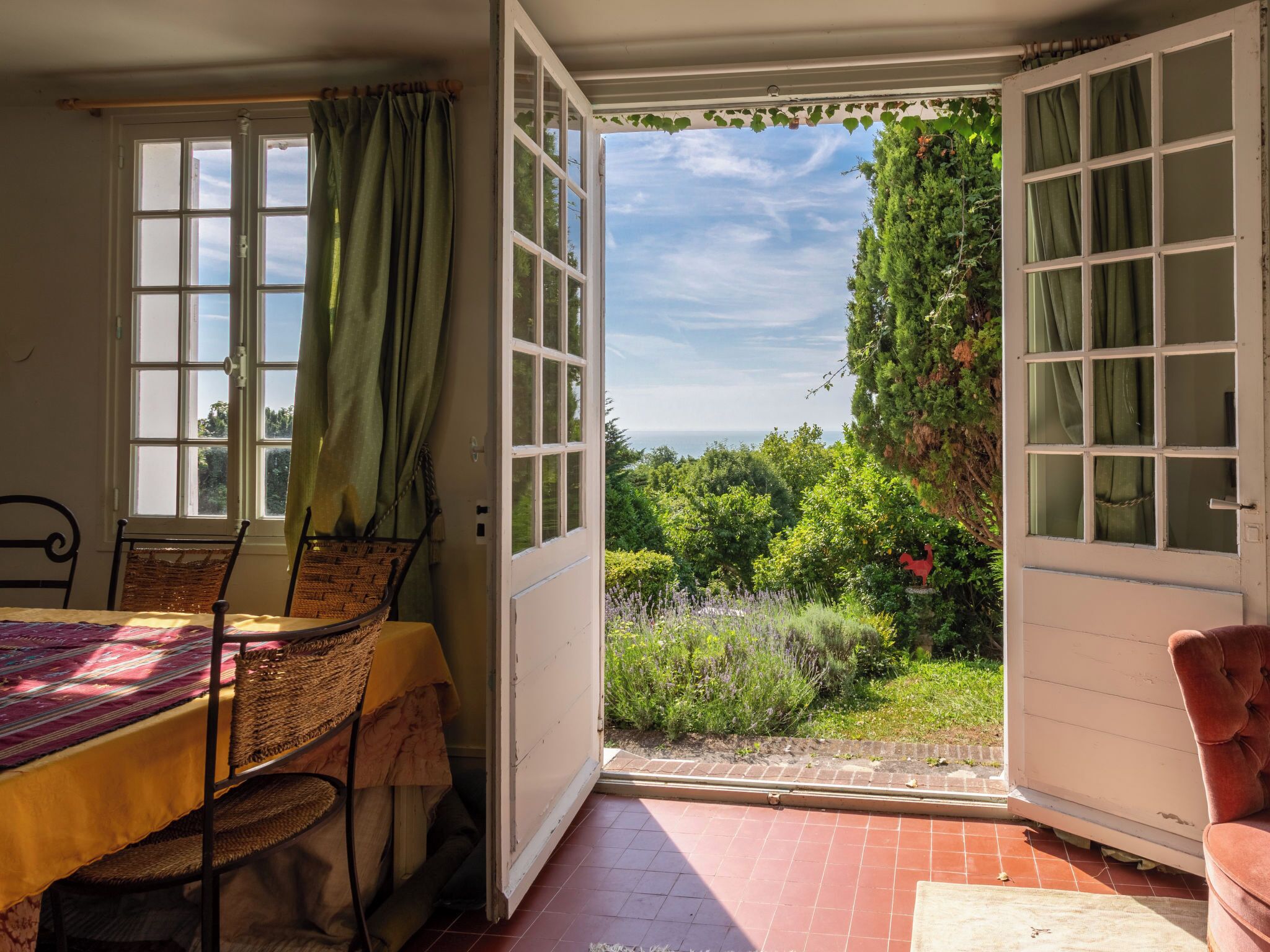 An open room with a table and chairs, a view of greenery and a red rooster statue outside.