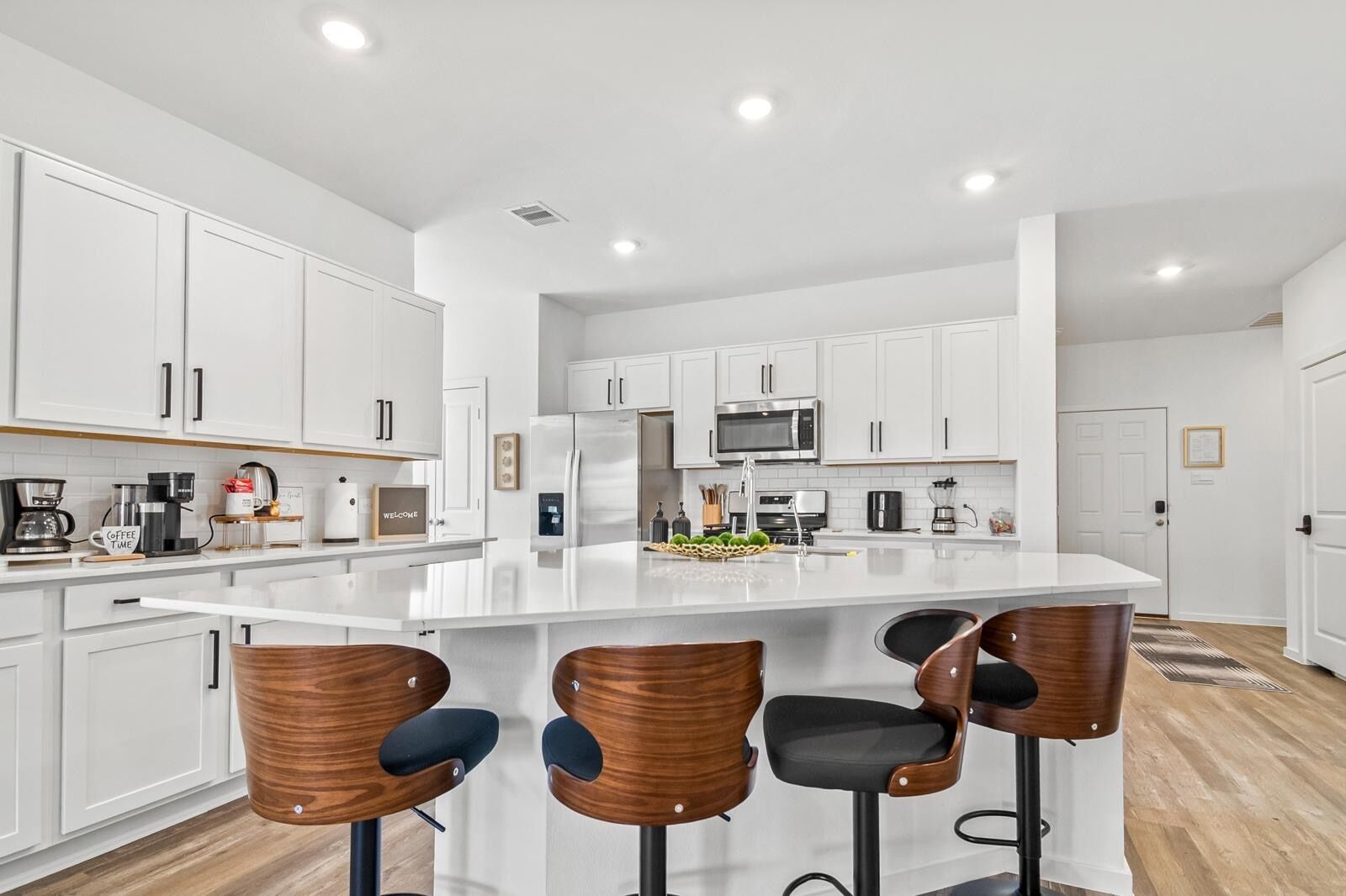 A modern kitchen with white cabinets, a large island with bar stools, stainless steel appliances, and a coffee station.
