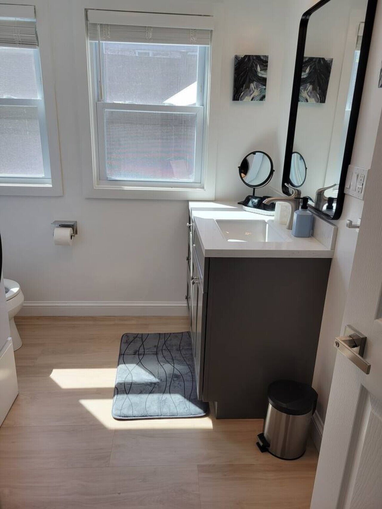 Un baño con un lavamanos blanco, dos espejos, un perchero y un cubo de basura.
