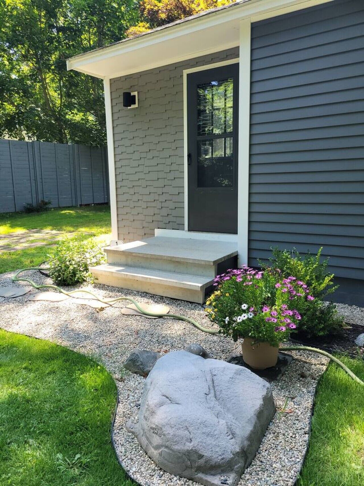 Una casa con fachada gris, una puerta principal, un pequeño porche con escalones, un jardín con una roca y una maceta con flores rosadas.