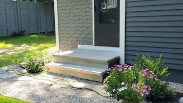 Una casa con fachada gris, una puerta principal, un pequeño porche con escalones, un jardín con una roca y una maceta con flores rosadas.