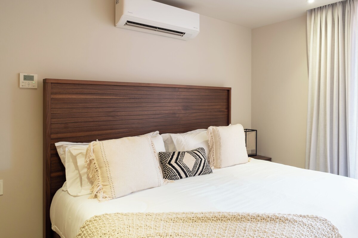 A hotel room with a wooden headboard, a bed with white bedding, a throw blanket, and a wall-mounted air conditioner.