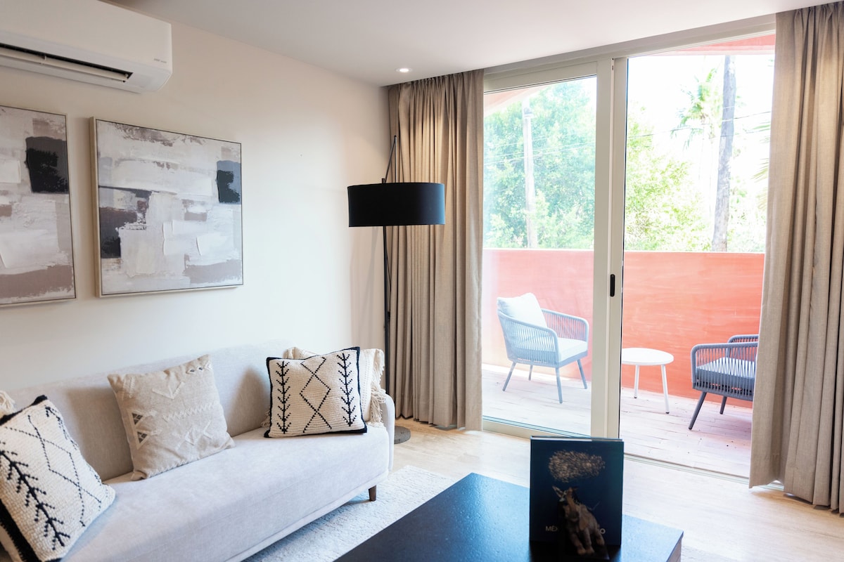 A modern hotel room with a sofa, a floor lamp, and a view of an outdoor patio with chairs.