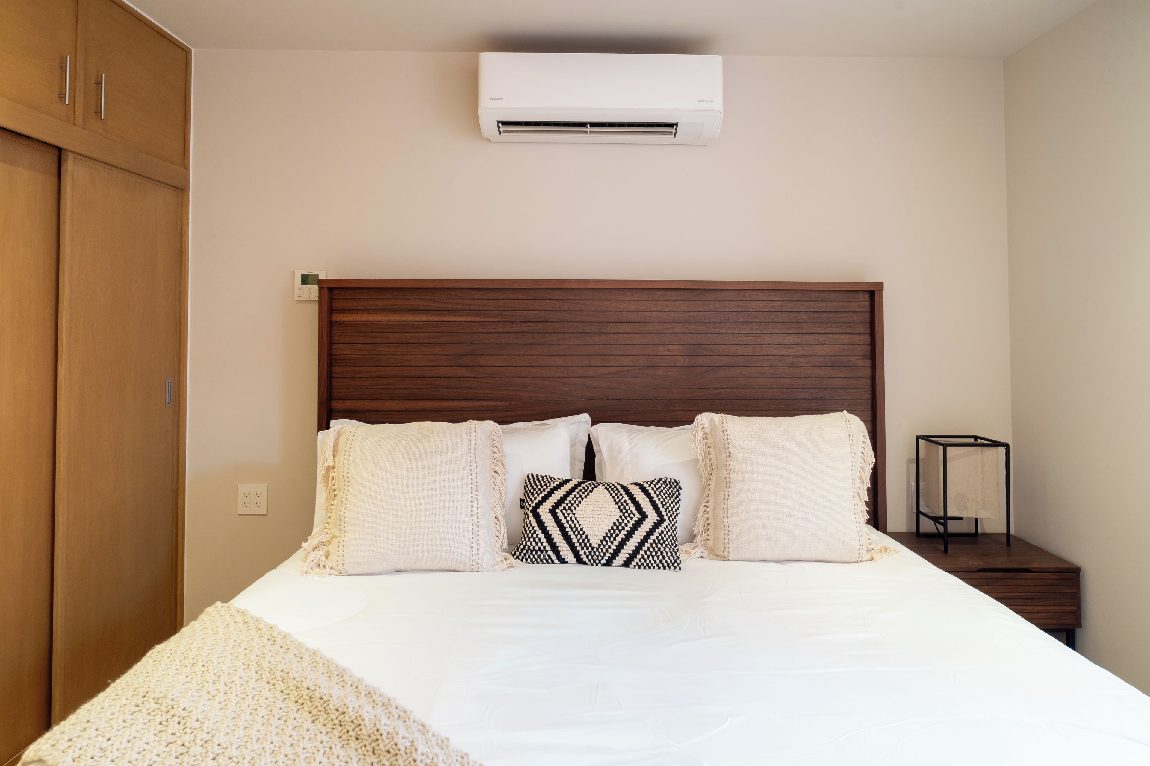 Une chambre à coucher avec une tête de lit en bois, un lit avec une literie blanche, deux coussins à motifs, une table de chevet et un appareil de climatisation.