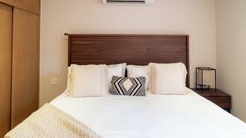 Une chambre à coucher avec une tête de lit en bois, un lit avec une literie blanche, deux coussins à motifs, une table de chevet et un appareil de climatisation.