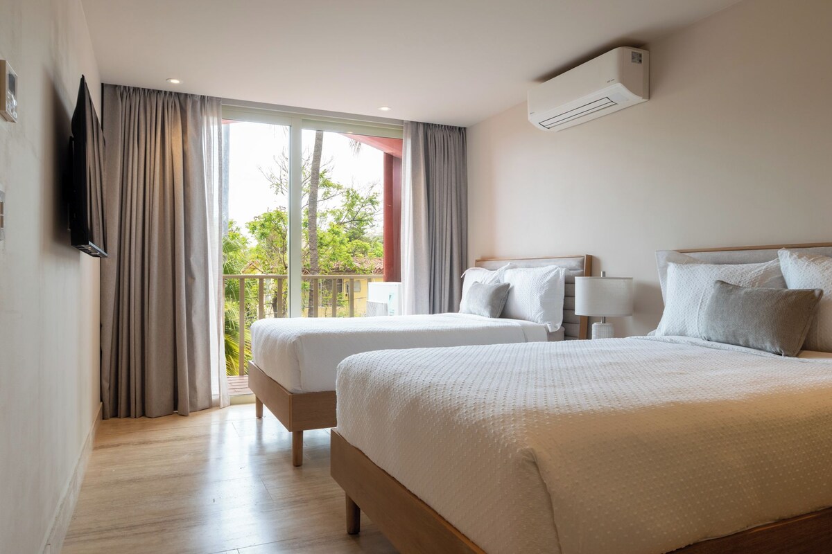 A hotel room with two beds, a TV, a balcony with curtains, and an air conditioning unit.