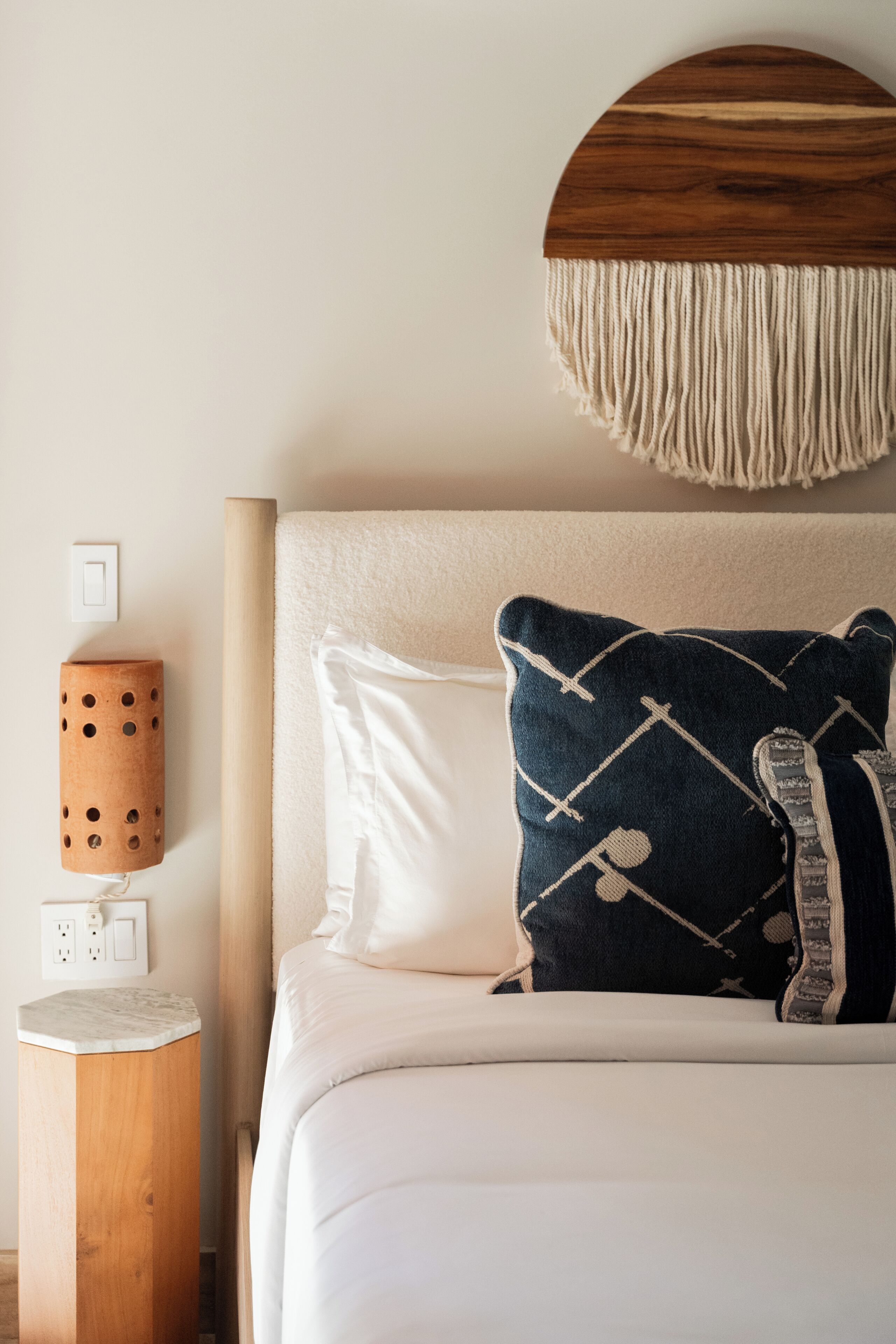 A neatly made bed with a white and blue patterned pillow, a wooden bedside table with a power outlet, and a wall-mounted switch.