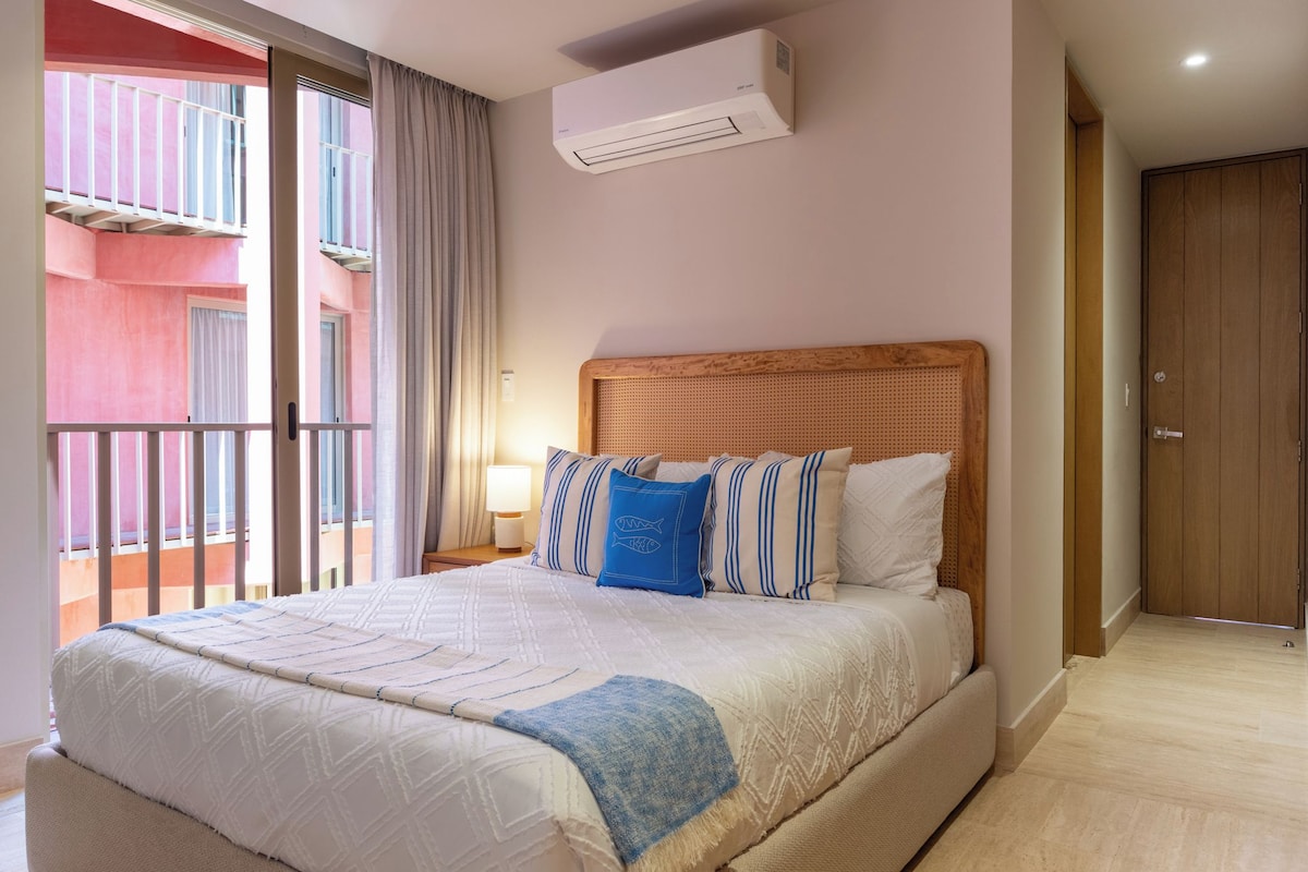 A hotel room with a bed, a nightstand, a lamp, an air conditioning unit, a balcony, and a wooden door.