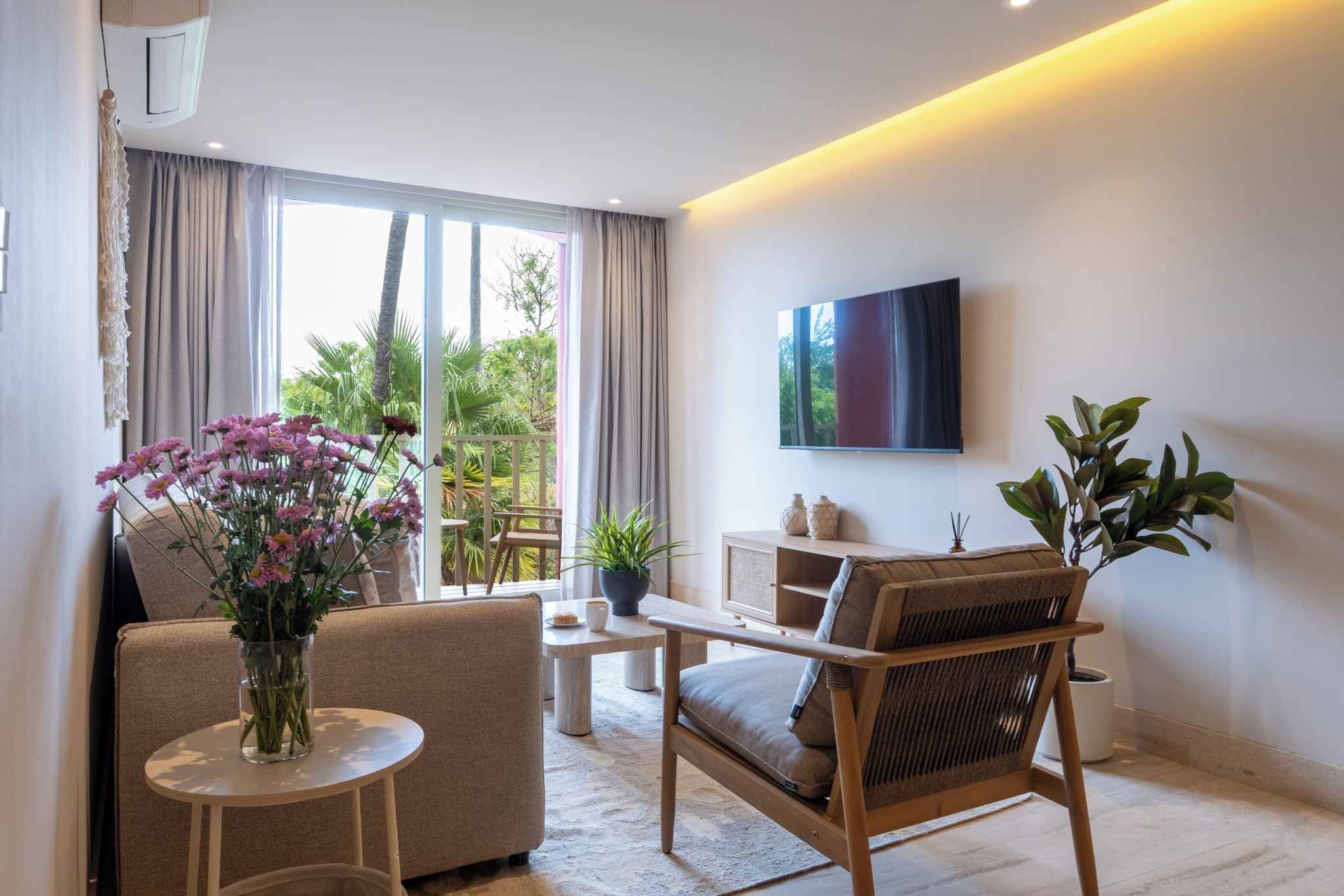 A modern living room with a sofa, a chair, a coffee table, and a TV mounted on the wall. There is a window with curtains, a plant on the windowsill, and a view of greenery outside.