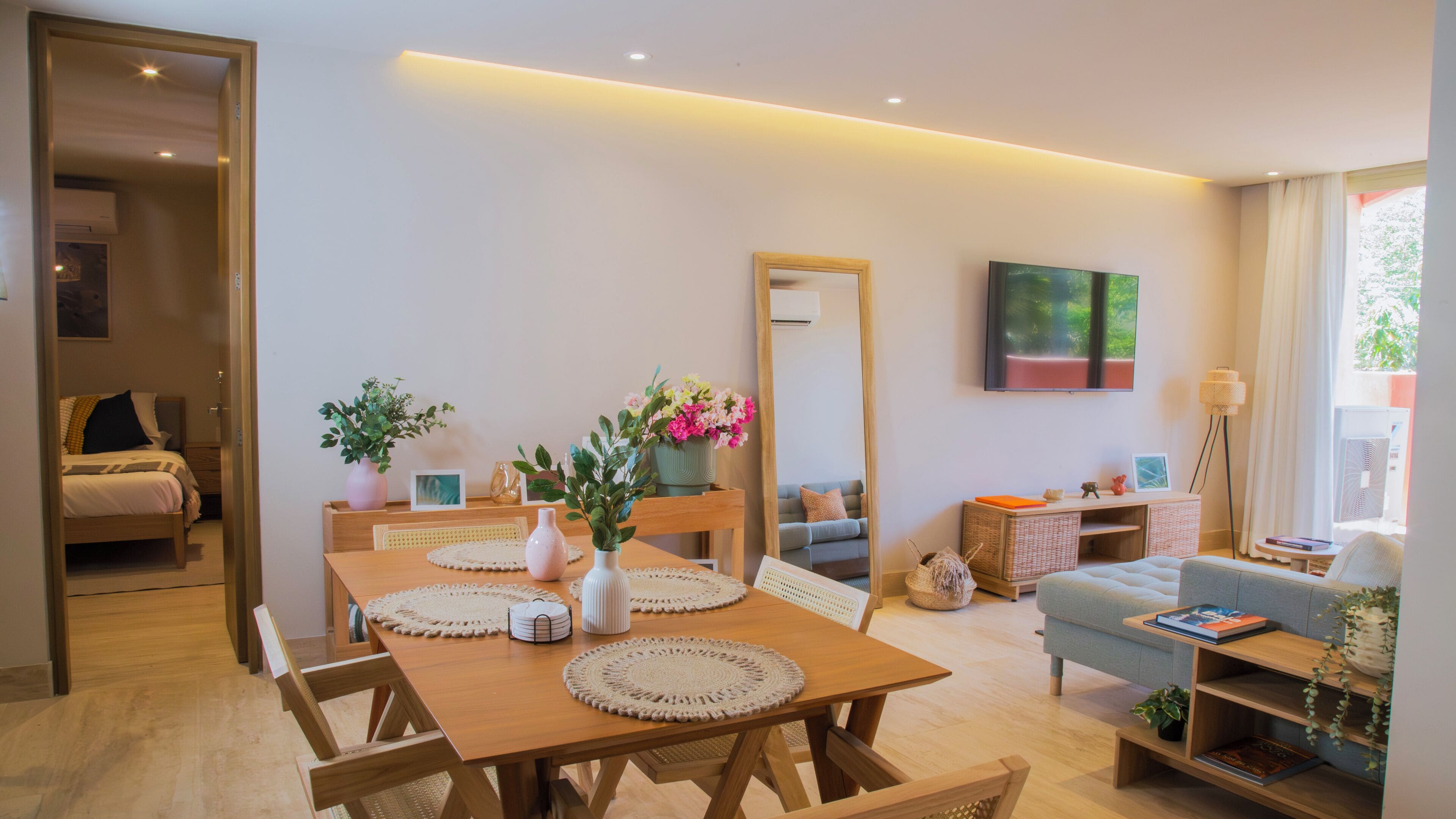 A modern living room with a wooden dining table, chairs, a sofa, a TV, and a large mirror.
