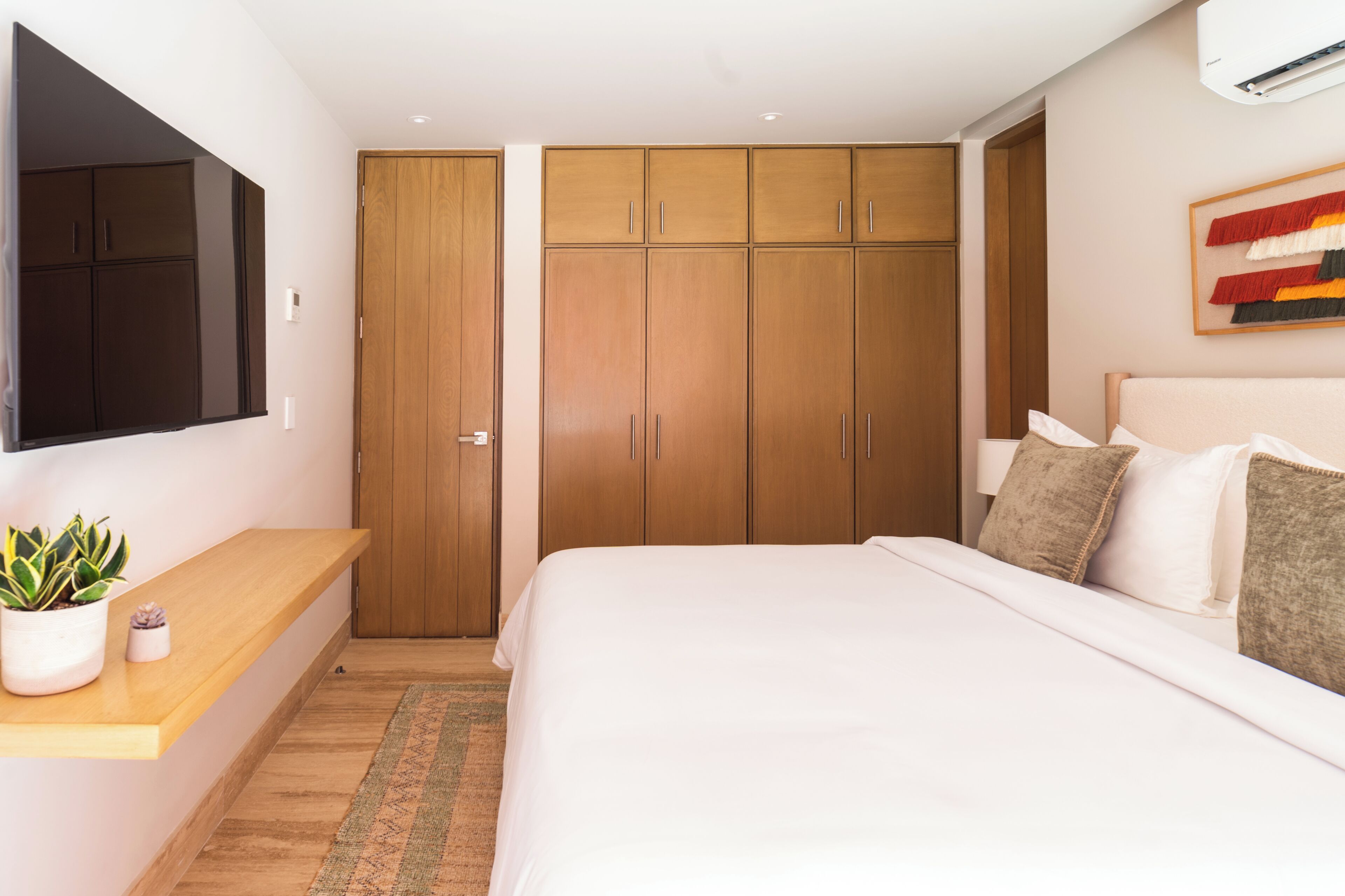 A bedroom with a bed, a TV mounted on the wall, a wardrobe, and a small plant on a shelf.