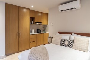 A bedroom with a bed, pillows, a bedside table, a small kitchen area, and a wall-mounted air conditioner.