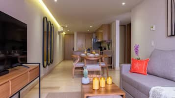 Una sala de estar moderna con un televisor de pantalla plana, una mesa de centro de madera con velas, un sofá gris con un cojín rojo, y una zona de comedor con sillas y una mesa de madera.