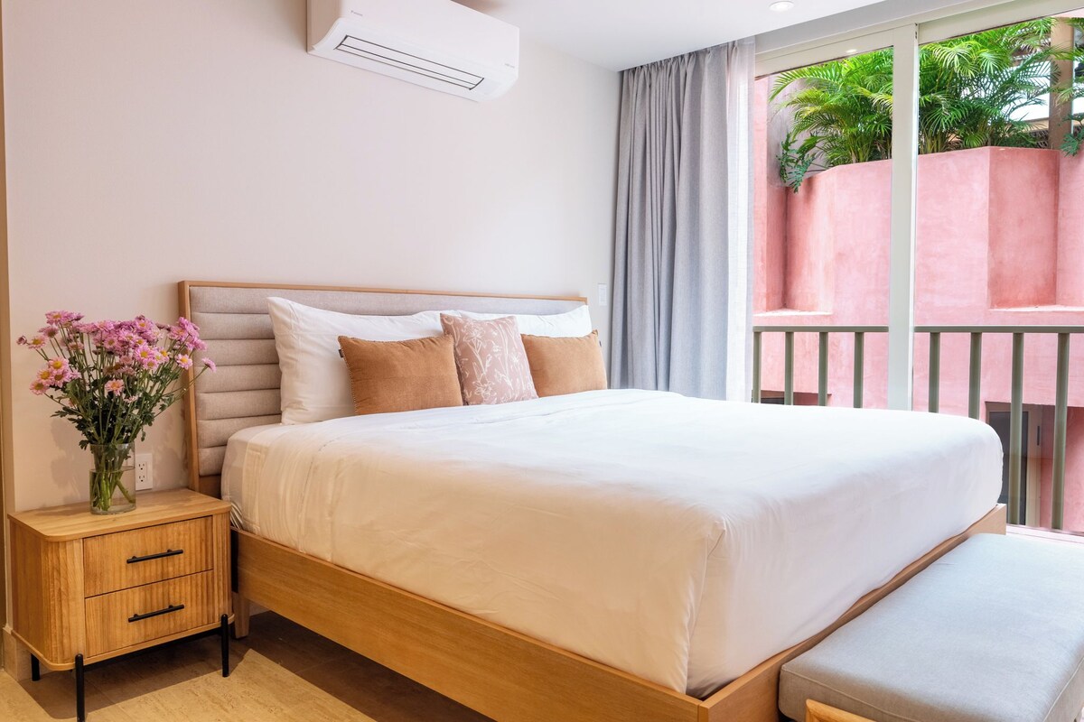 A hotel room with a bed, bedside table, and a view of a balcony with plants.