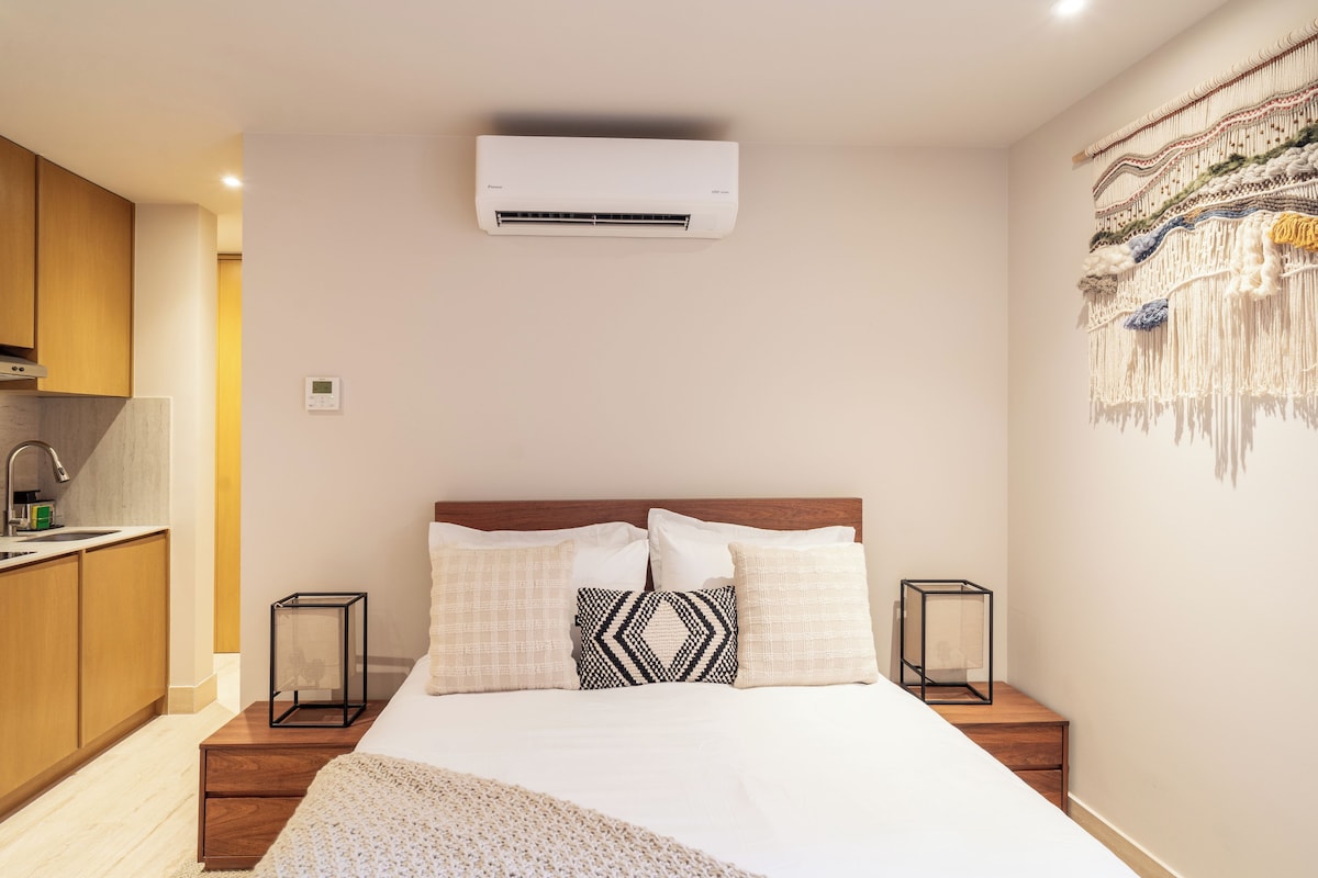 A hotel room with a bed, bedside tables, a wall-mounted air conditioner, a kitchen area, and a decorative wall hanging.