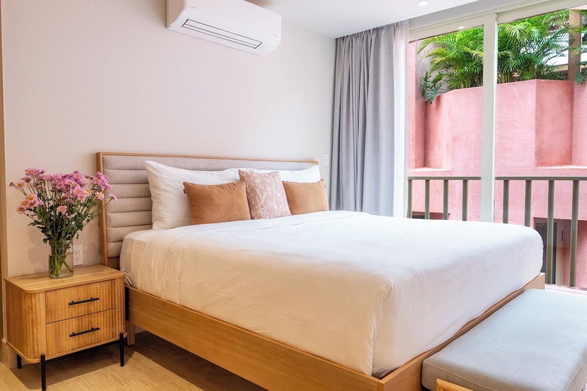 Habitación de hotel con cama, mesita de noche y vistas a un balcón con plantas.