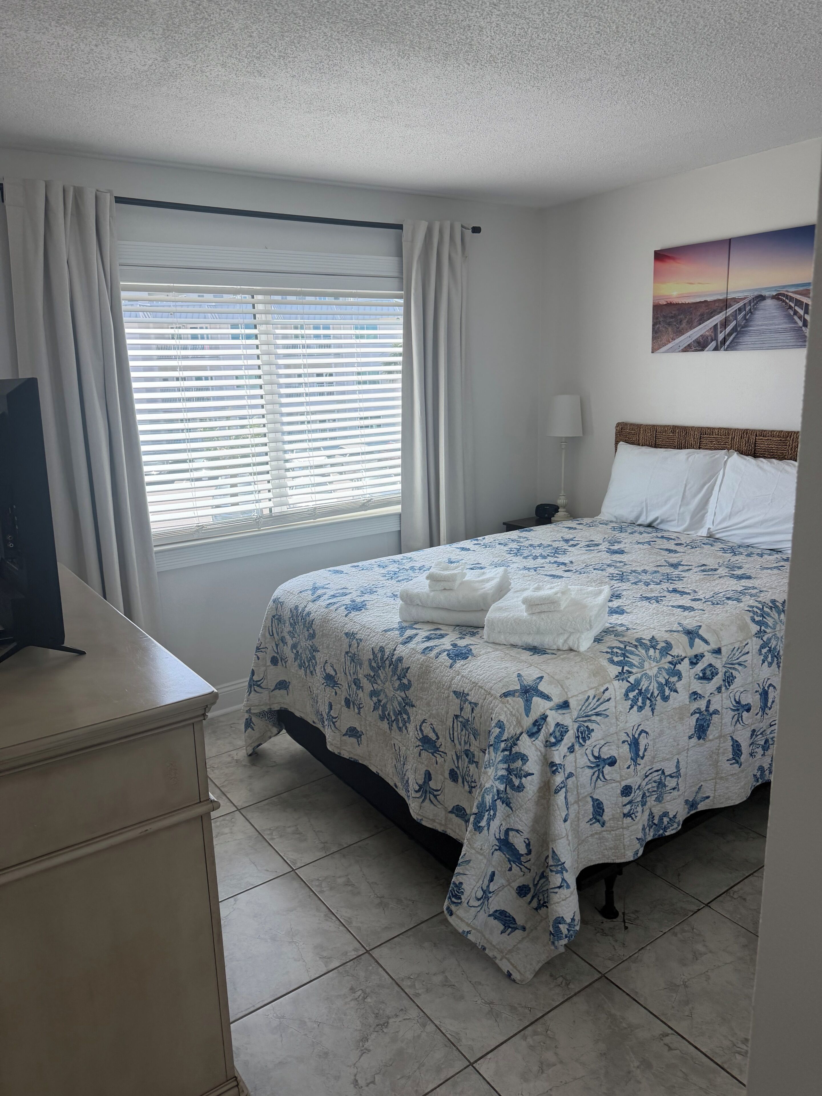 A hotel room with a bed, a desk, a window with curtains, a wall-mounted TV, and a painting.