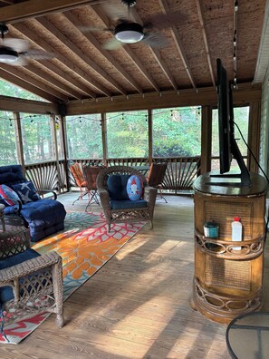 A covered outdoor seating area with wicker furniture, a patterned rug, and a wooden pillar with a television.
