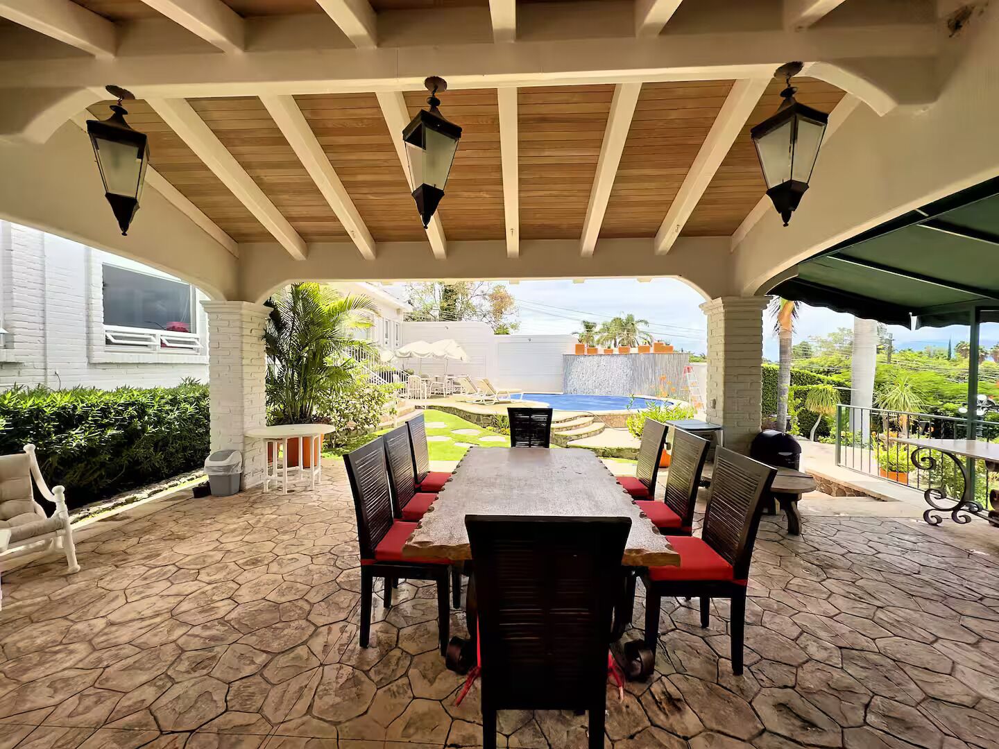 Un porche cubierto con piso de piedra, una mesa con sillas y vistas a una piscina y vegetación.