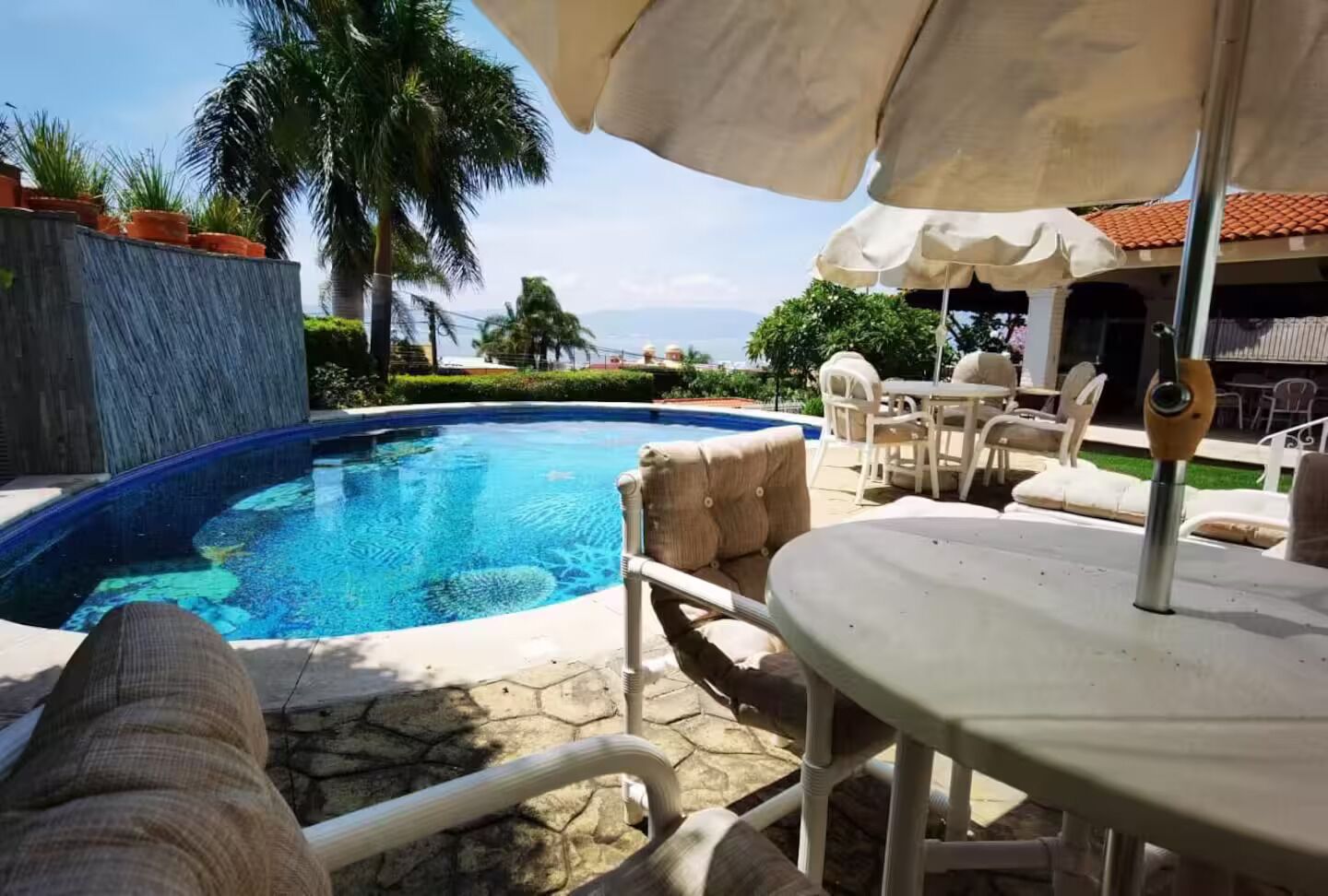 A swimming pool with a stone border, surrounded by lounge chairs and umbrellas, and palm trees in the background.