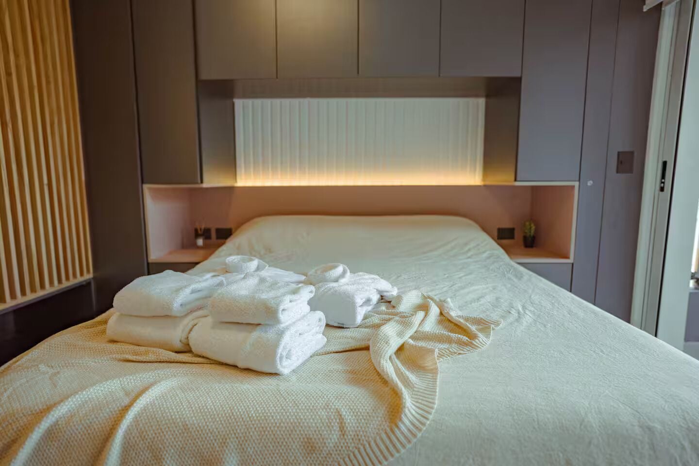 A neatly made bed with folded towels, a bedside table, and a wall-mounted shelf.