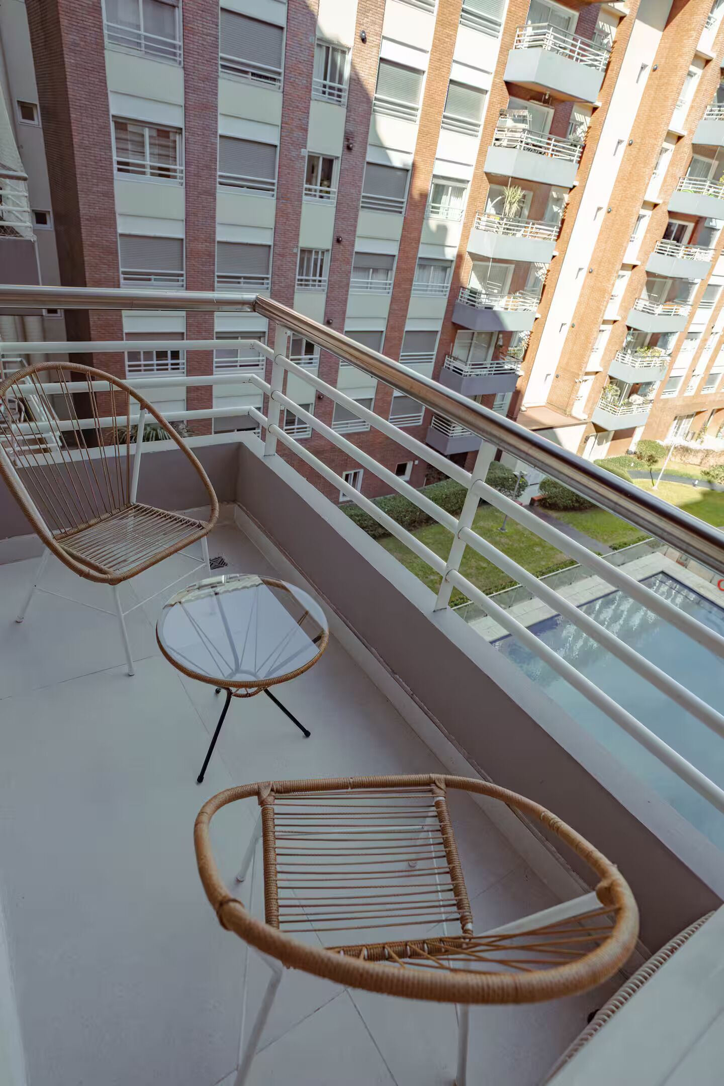 Un balcón con muebles de mimbre, una mesa de vidrio y vista a un edificio de varios pisos.