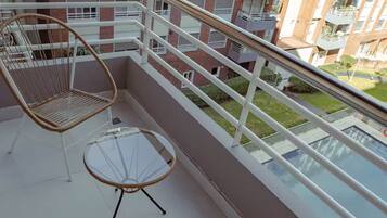 Un balcón con muebles de mimbre, una mesa de vidrio y vista a un edificio de varios pisos.