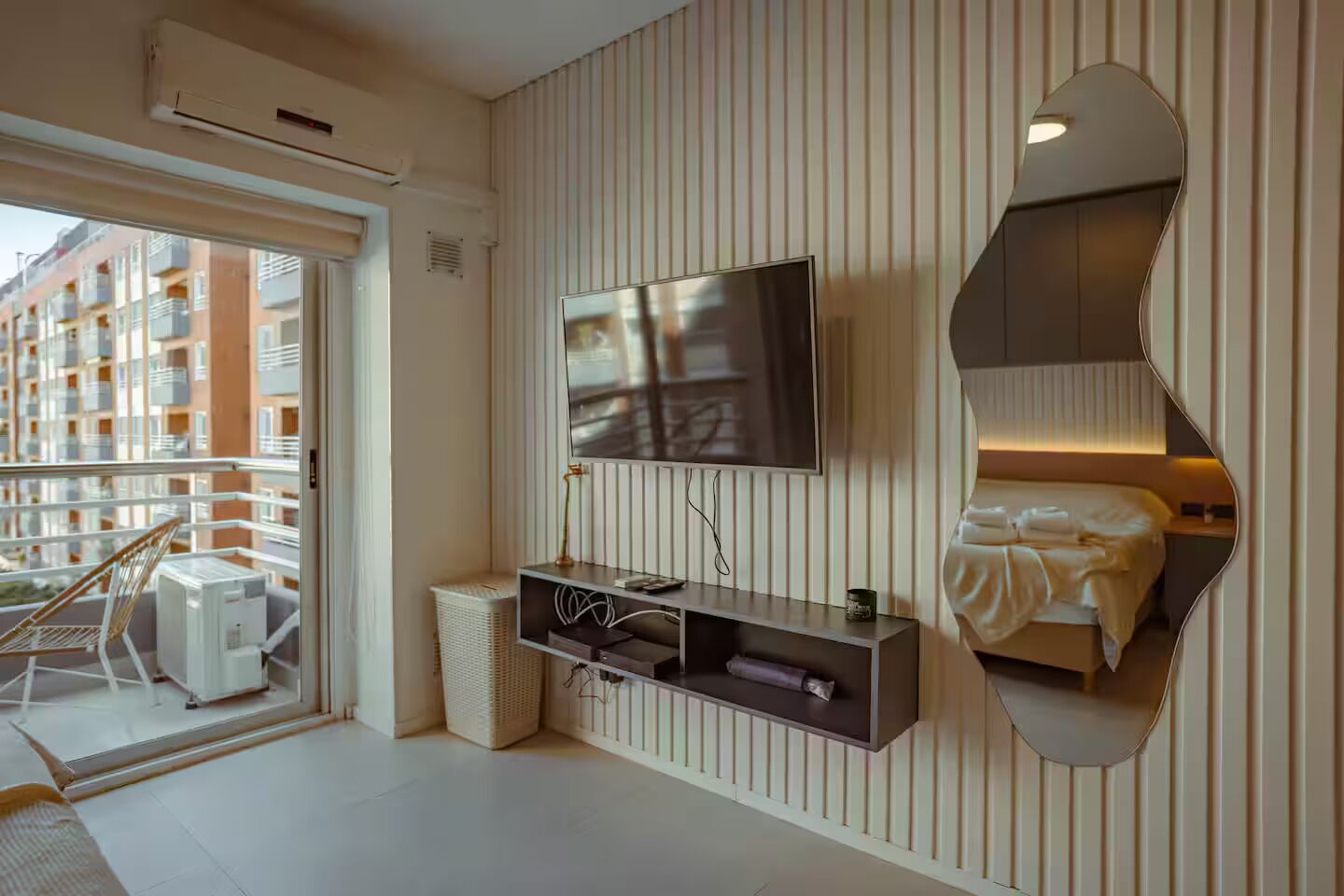 A modern hotel room with a large TV, a mirrored wall, a balcony with a chair, and a visible city view.