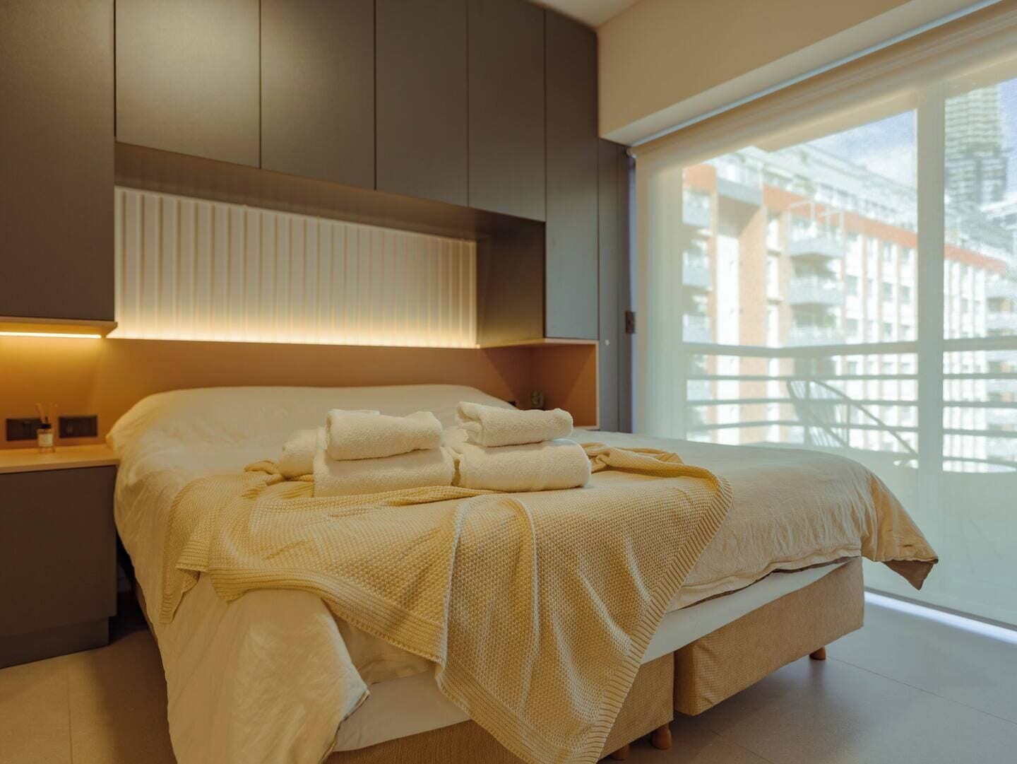 A modern bedroom with a large bed, a bedside table, and a view of the city through a large window.