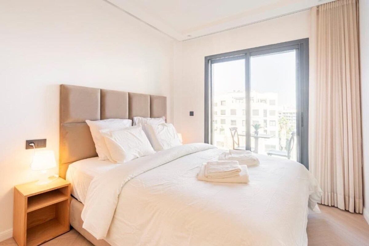 Ein Hotelzimmer mit einem Bett, einem Nachttisch, einem Fenster mit Vorhängen und Blick auf Gebäude.