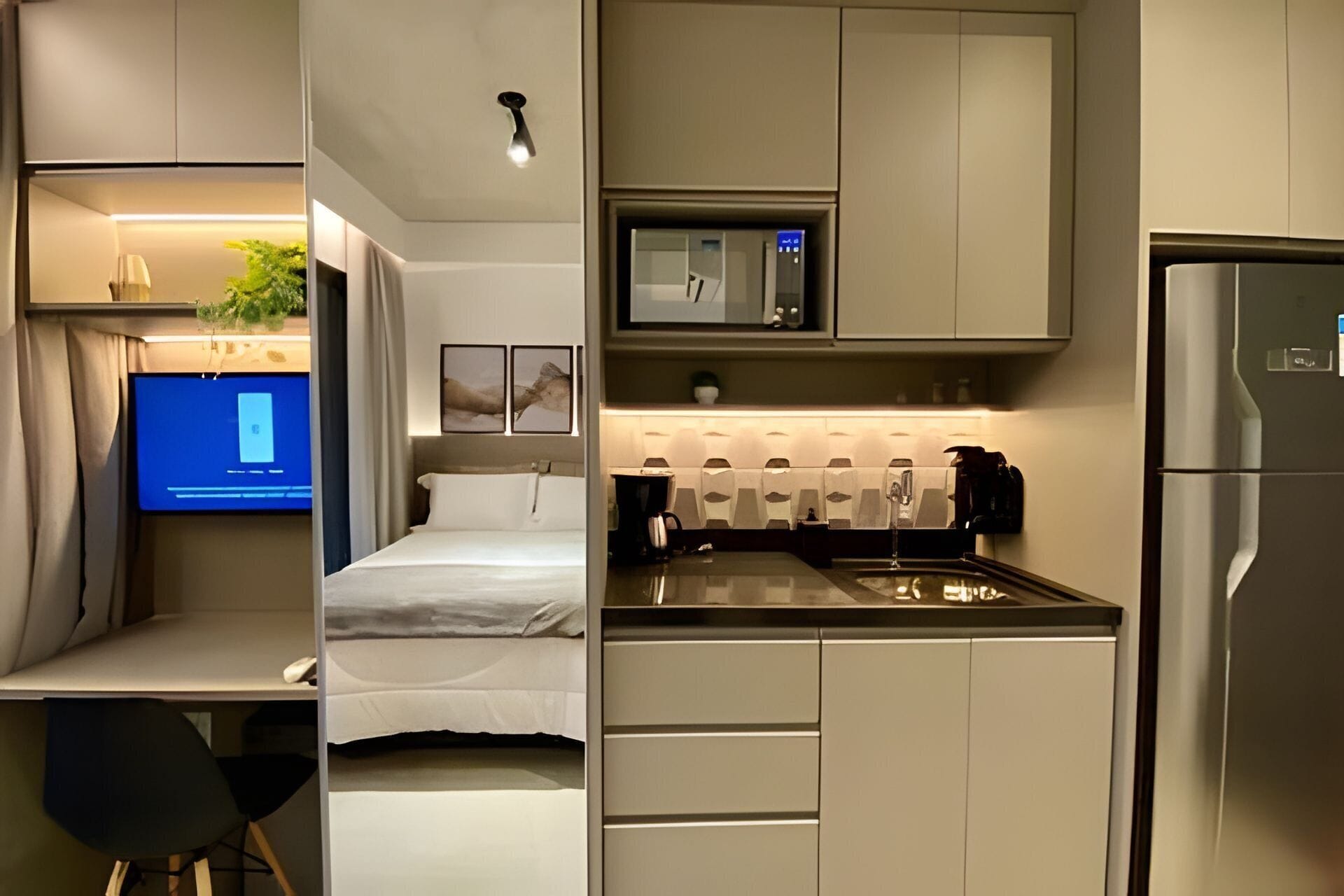 Ein kleines Hotelzimmer mit Bett, einem Schreibtisch, einer Küchenzeile inklusive Kühlschrank, Mikrowelle und Kaffeemaschine.