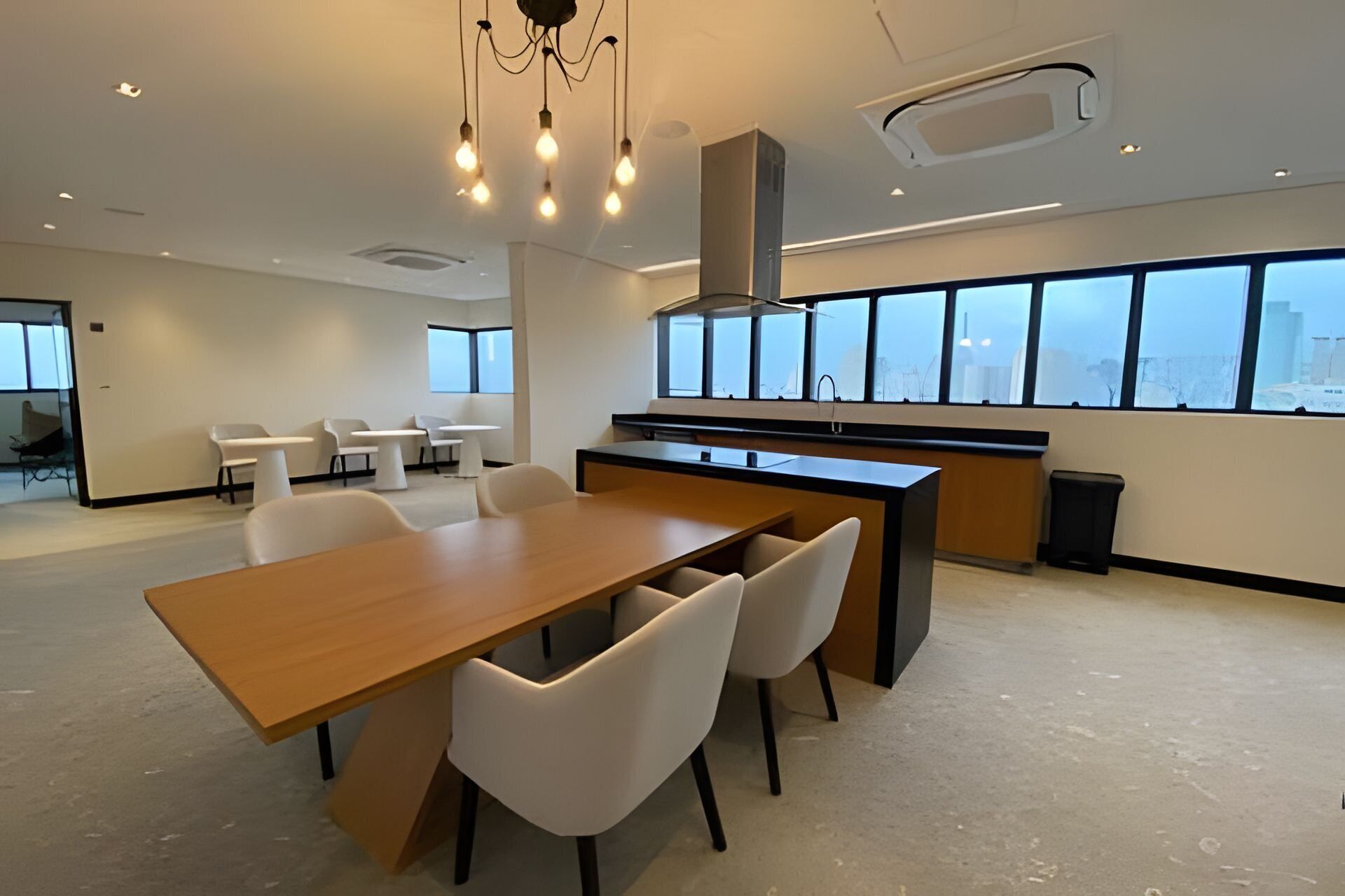 Un espace de bureau moderne doté d’une longue table en bois, de chaises blanches, d’un coin cuisine et de grandes fenêtres offrant une vue sur la ville.