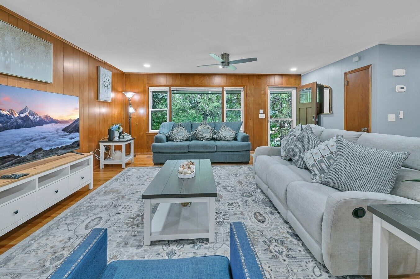 Ein Wohnzimmer mit Sofa, Couchtisch und Fernseher, Holzwandverkleidung und großen Fenstern.