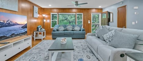 Una sala de estar con sofá, mesa de centro y televisor, paredes de paneles de madera y ventanas amplias.