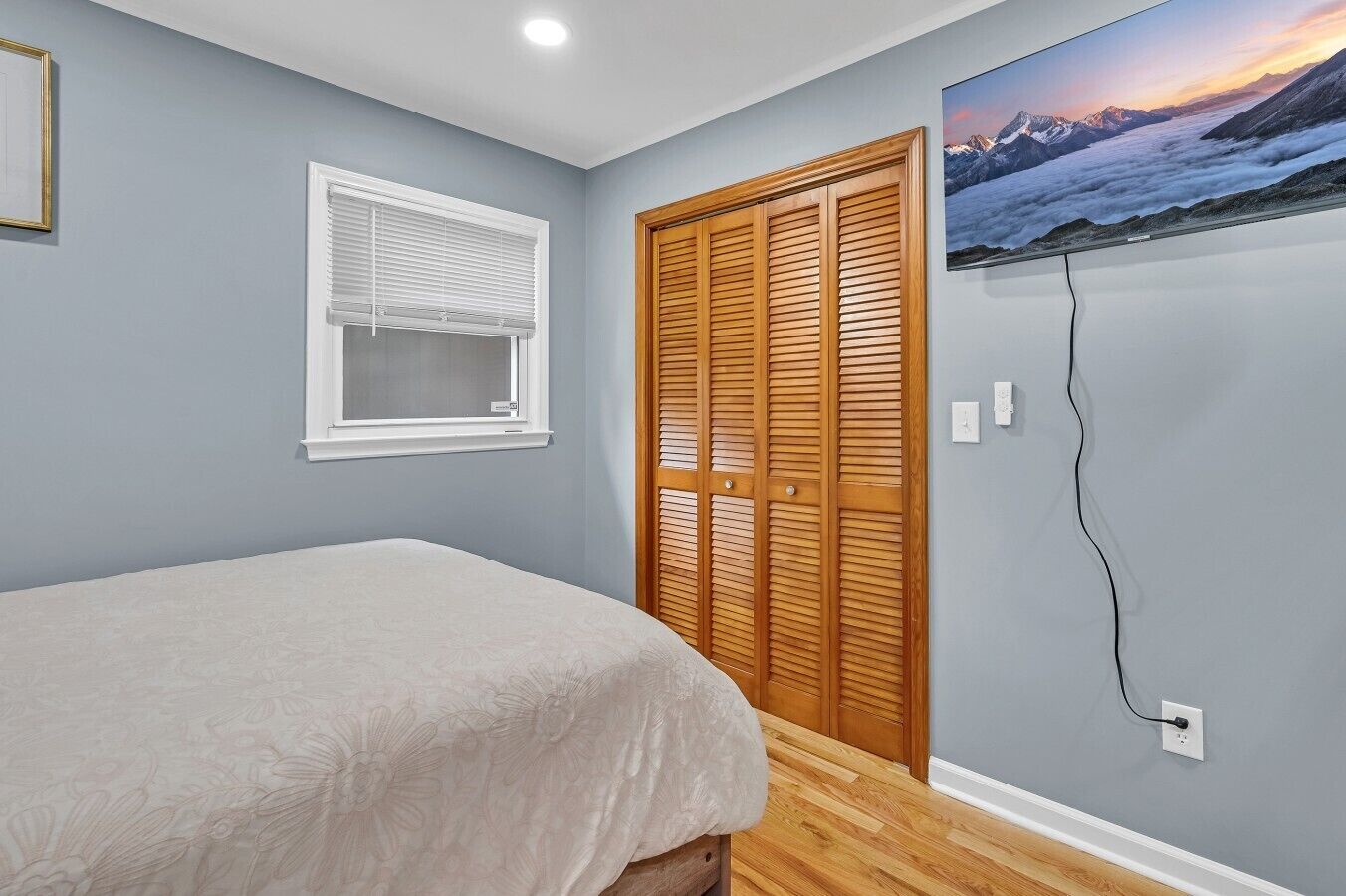 A bedroom with a bed, a window with blinds, a wooden closet, and a wall-mounted TV displaying a mountain landscape.