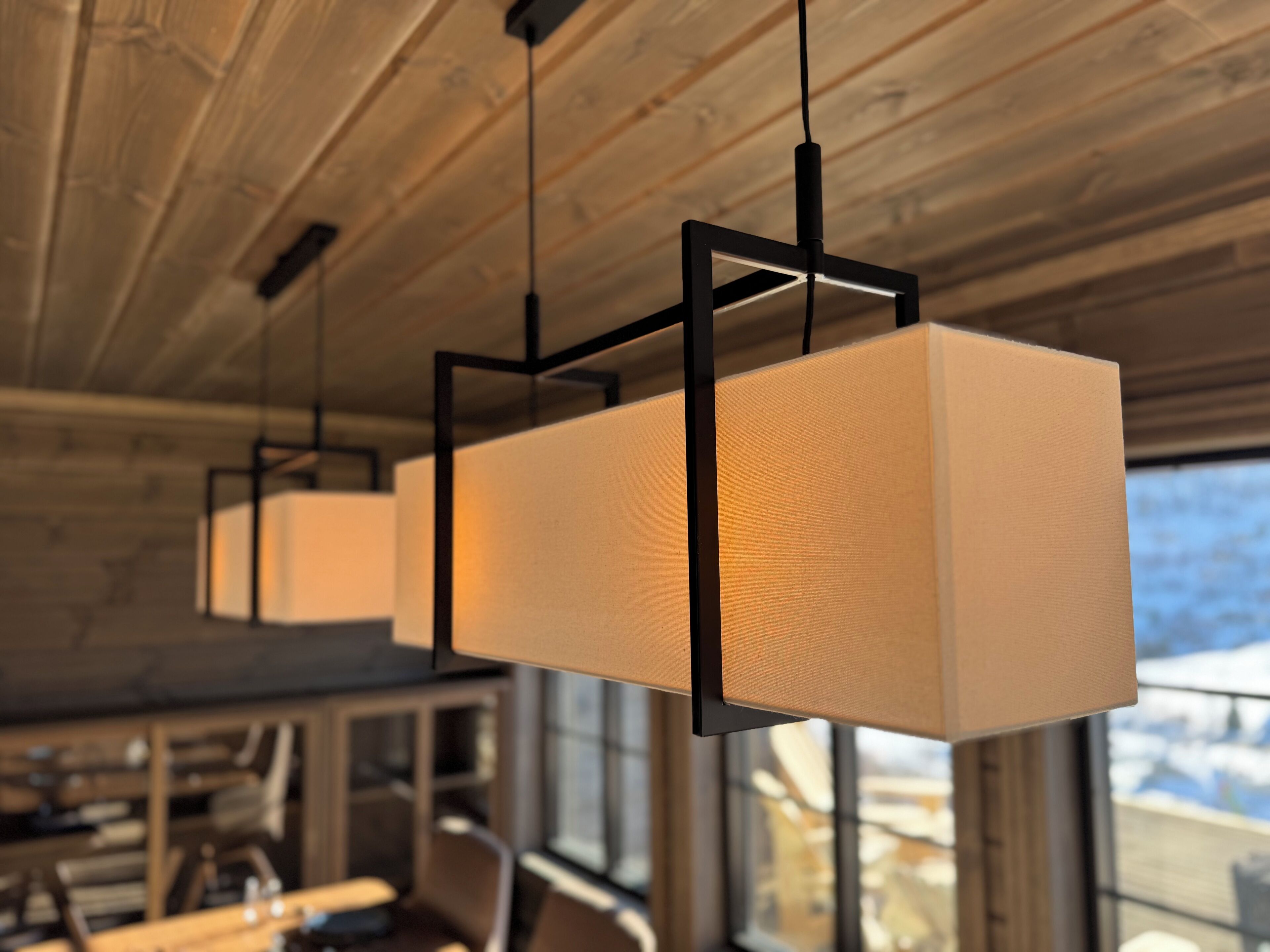 A modern interior with wooden ceiling beams, pendant lights with square shades, and a view through a window.
