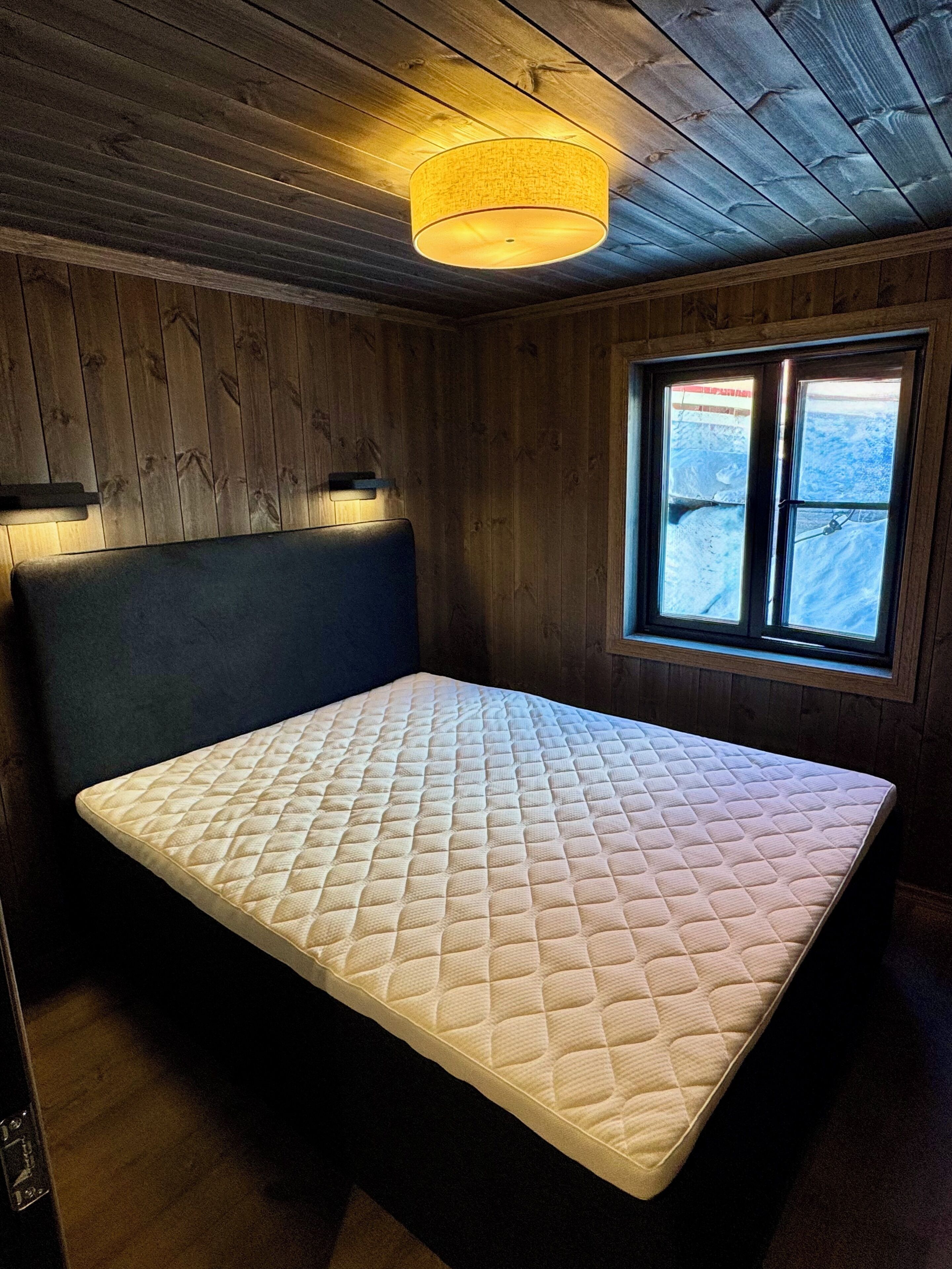 Una habitación con cama, paredes de madera, ventana con vistas y una lámpara en el techo.