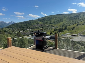 A wooden deck with a barbecue grill overlooking a mountainous landscape with a village below.