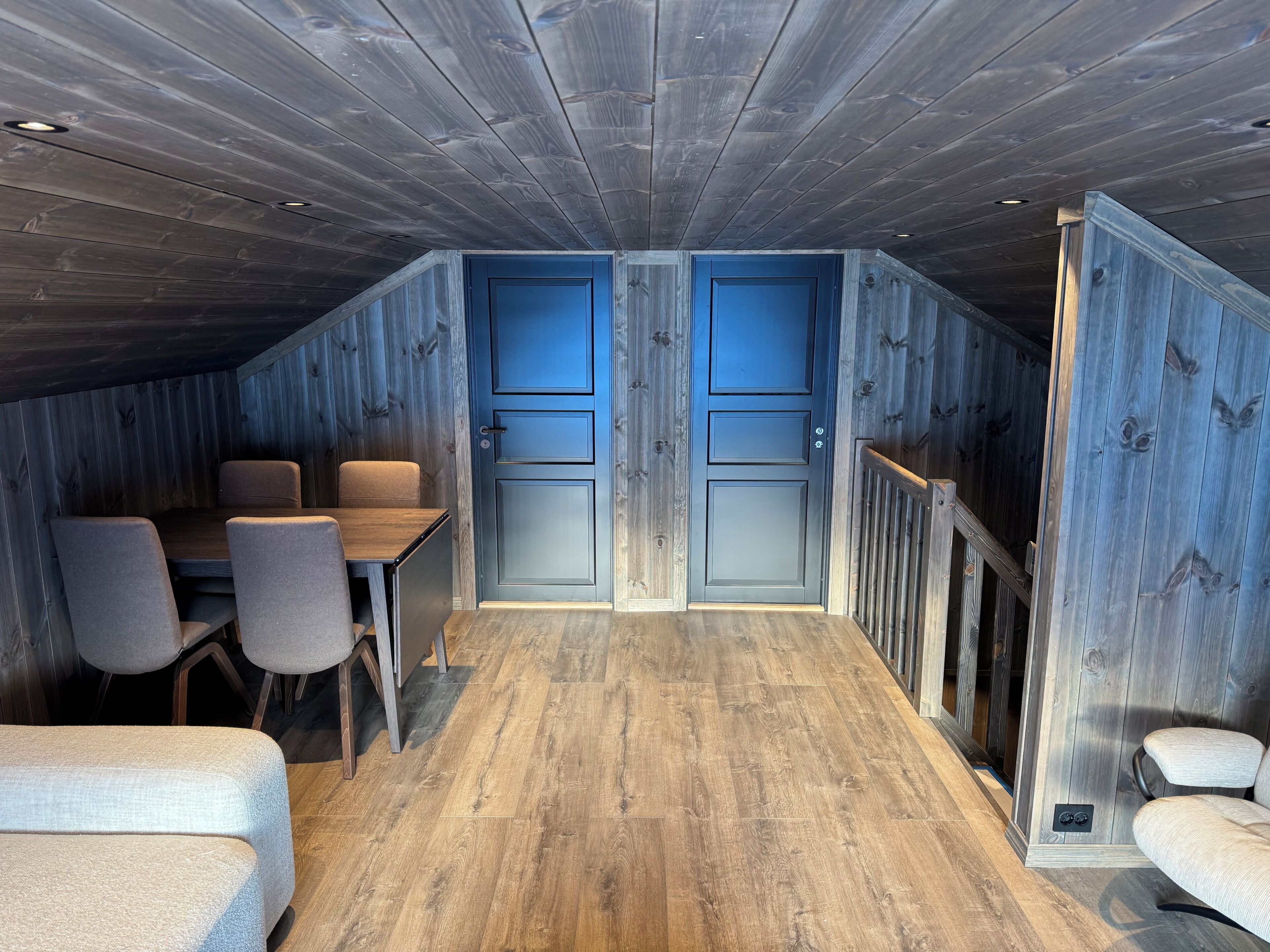 Une pièce avec un plafond en pente, un plancher en bois, une table à manger avec des chaises et deux portes.