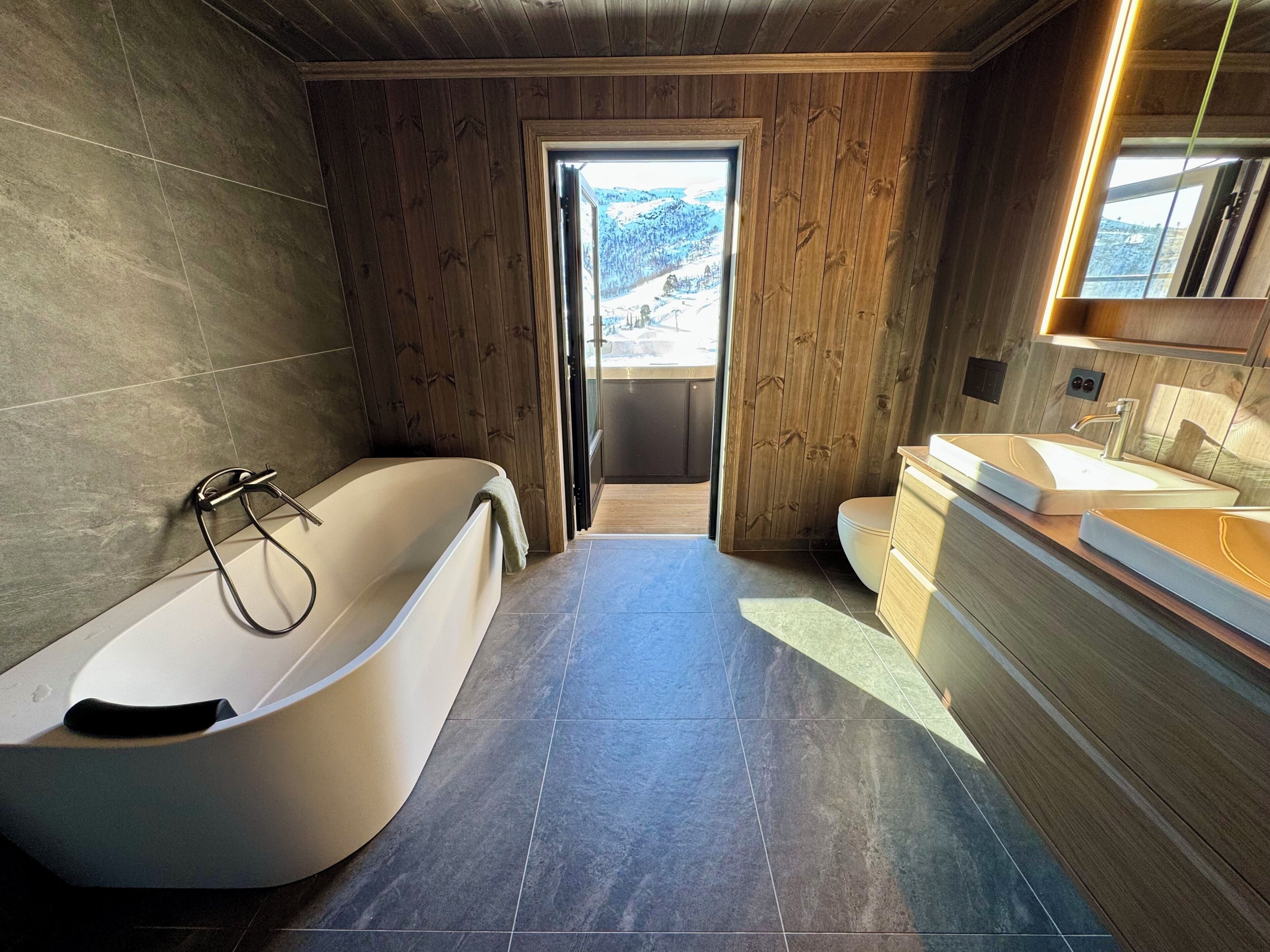 Un baño moderno con una bañera independiente, un tocador doble y vistas a un paisaje nevado a través de una puerta abierta.