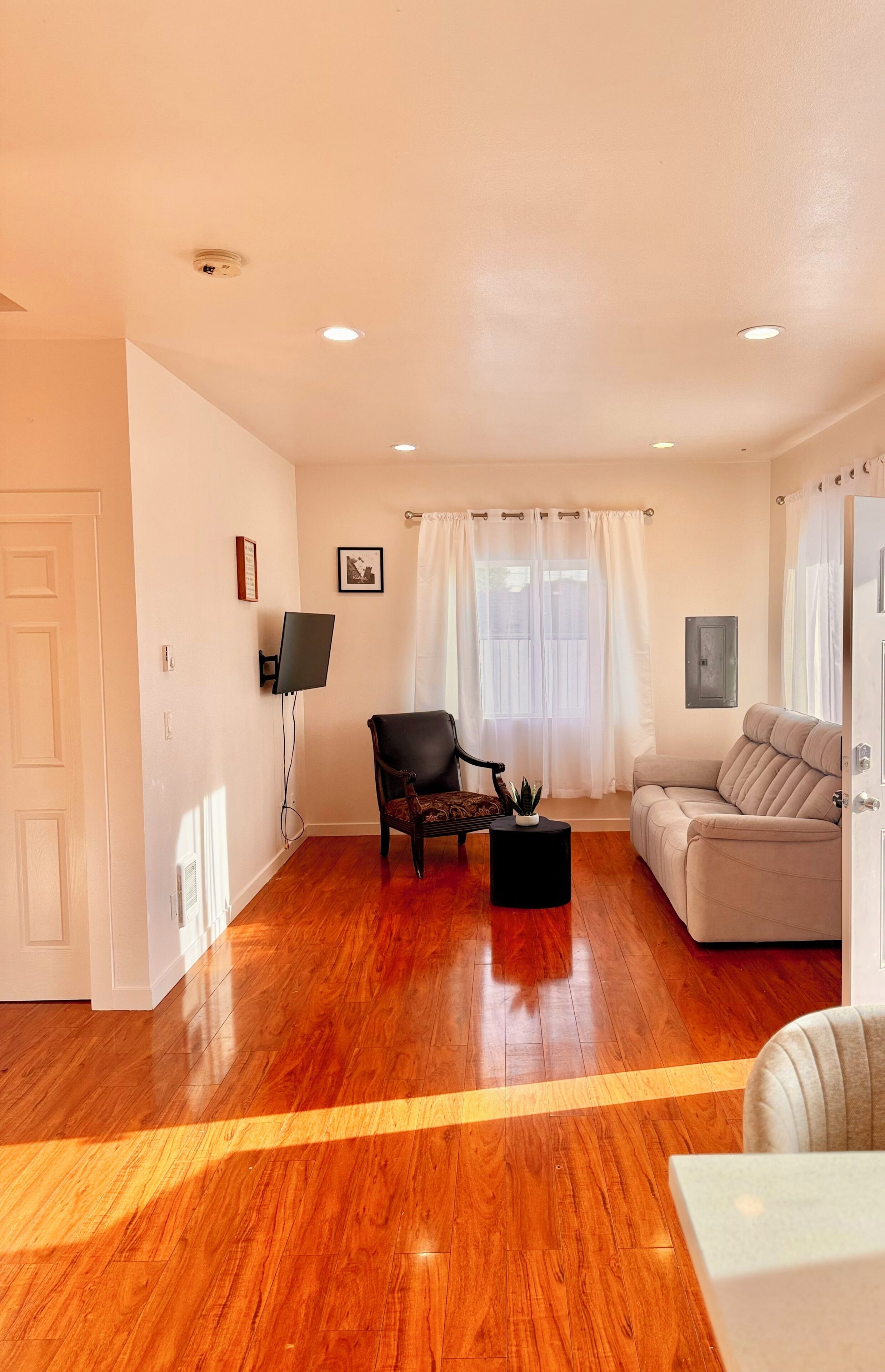 A living room with hardwood flooring, a black armchair, a beige sofa, a small black table, and a mounted flat-screen TV.
