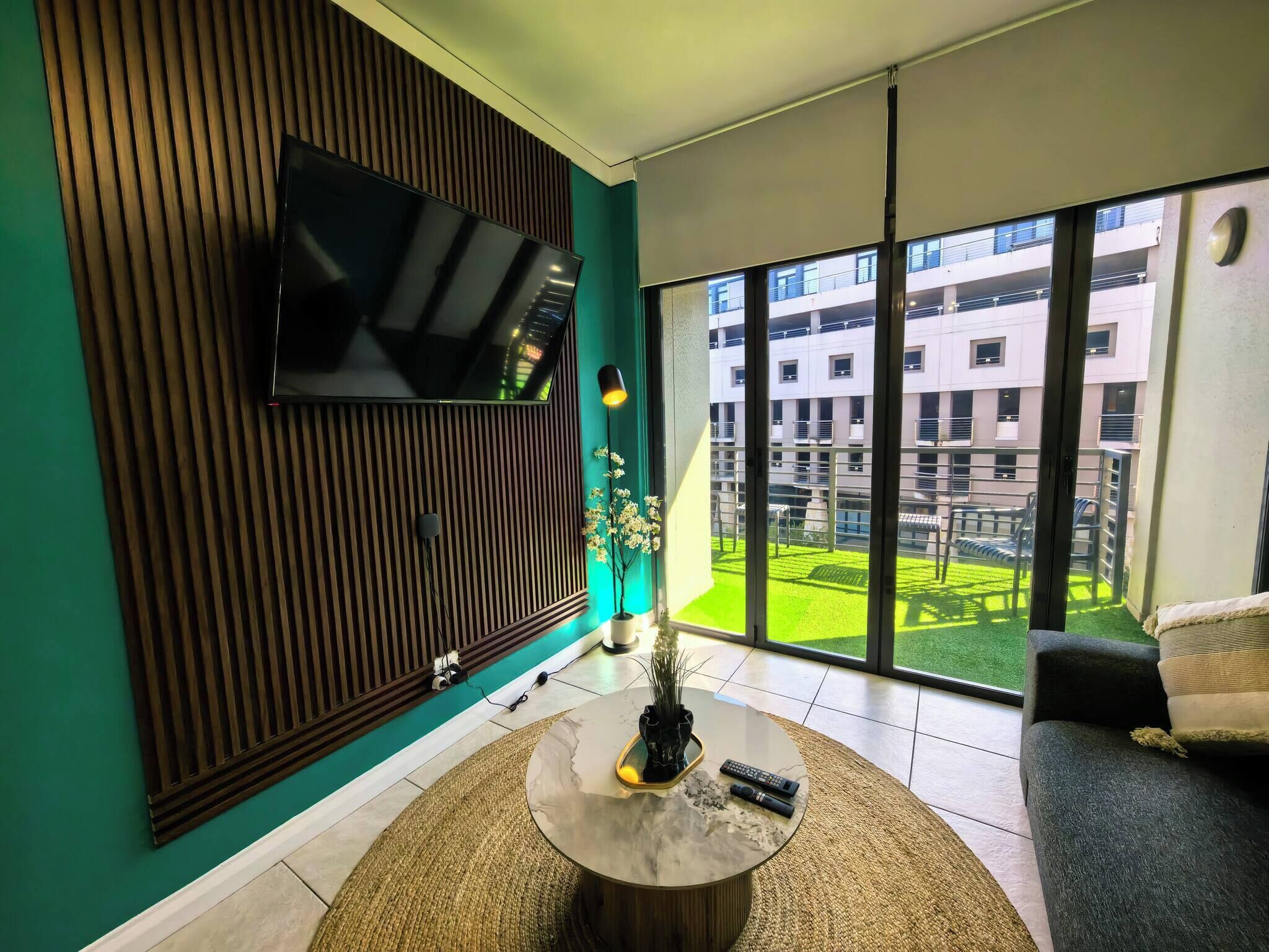 A modern living room with a flat-screen TV mounted on a wooden panel wall, a round coffee table with a vase and remote controls, a sofa, and a large window offering a view of a building and a green space.