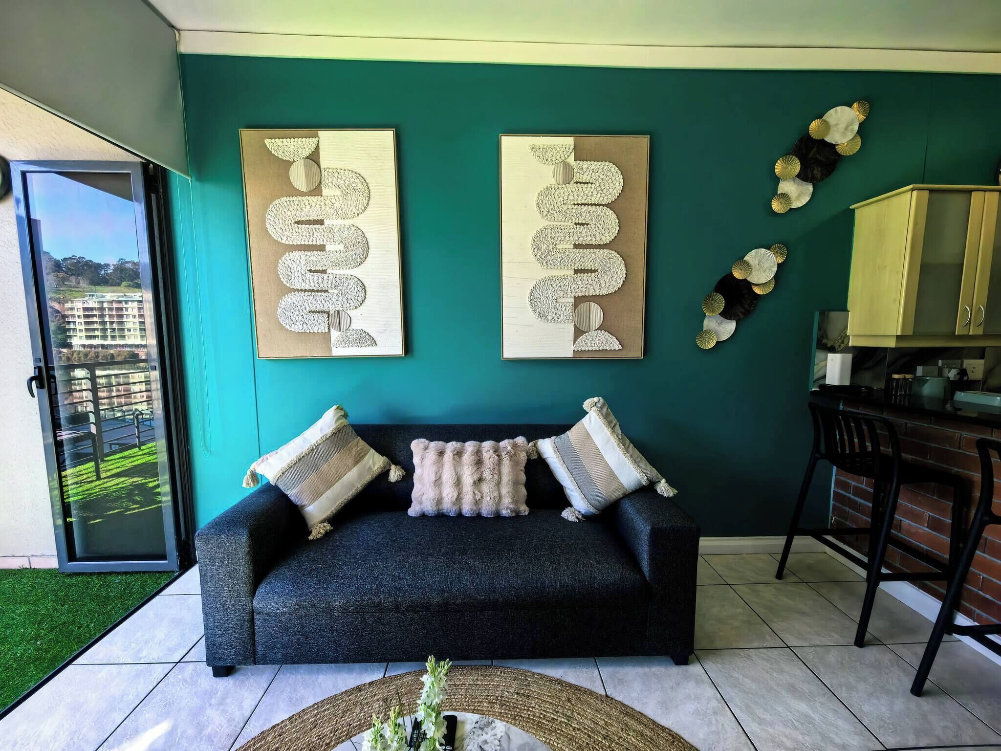 Een woonkamer met teal muren, een donkergrijze bank, twee abstracte kunstwerken aan de muur, en een keukentje met een geel kastje.