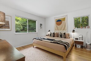 Un dormitorio con cama, mesitas de noche, lámpara, espejo y ventana con vistas a zonas verdes.
